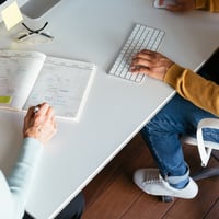 Colleagues collaborate at a workstation, one taking handwritten notes, one using their keyboard.