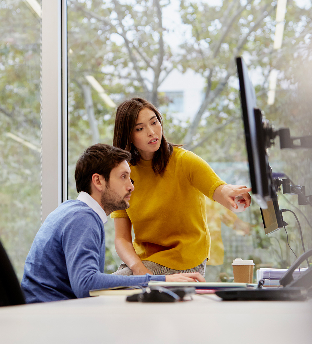 Two colleagues gather at one desk, referencing material on the computer monitor.