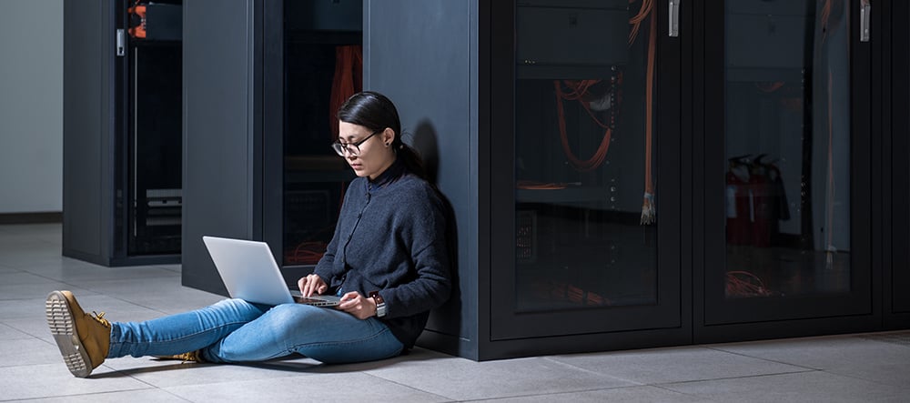 A young woman sitting on the floor against a server cabinet, typing on her laptop.