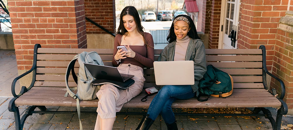 Two colleagues working remotely from an outdoor bench, looking at their laptops during conversation.