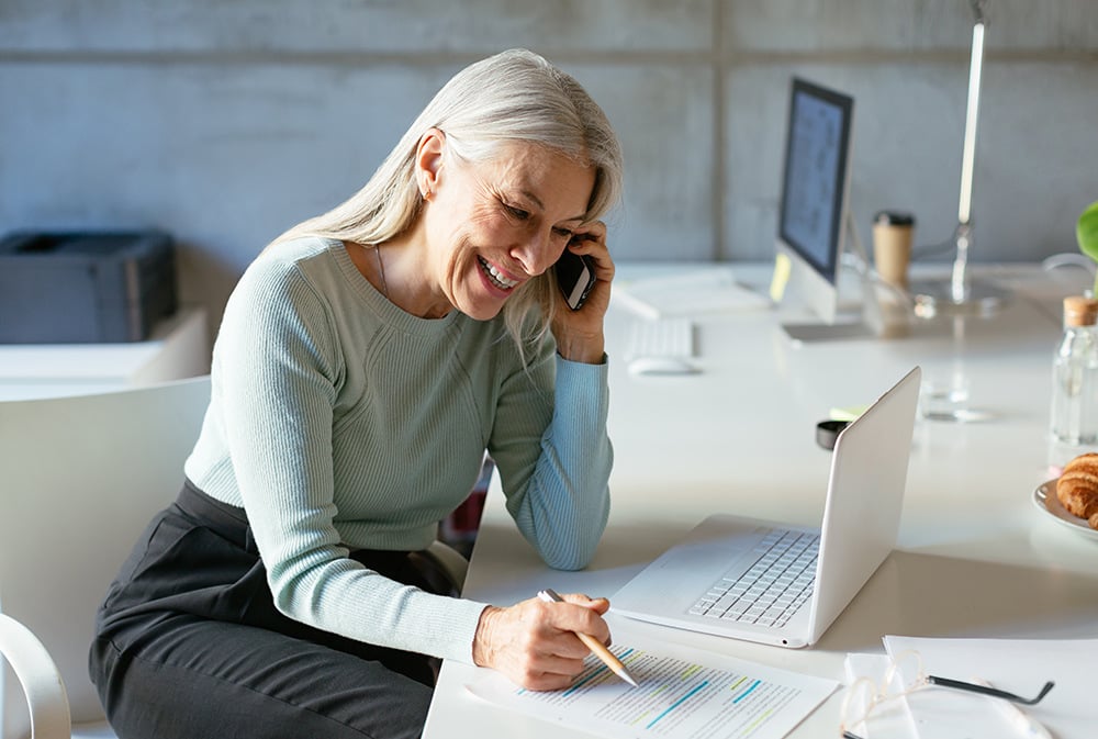 An office worker happily using her laptop, talking on the phone, and taking notes simultaneously. 