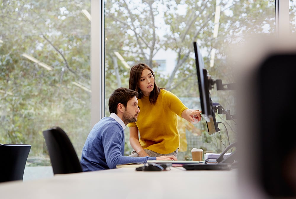 Two colleagues gather at one desk, referencing material on the computer monitor.