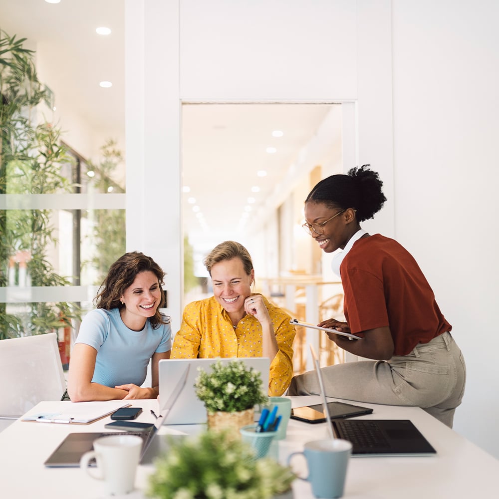 Three smiling professional employees look at a shared laptop screen.