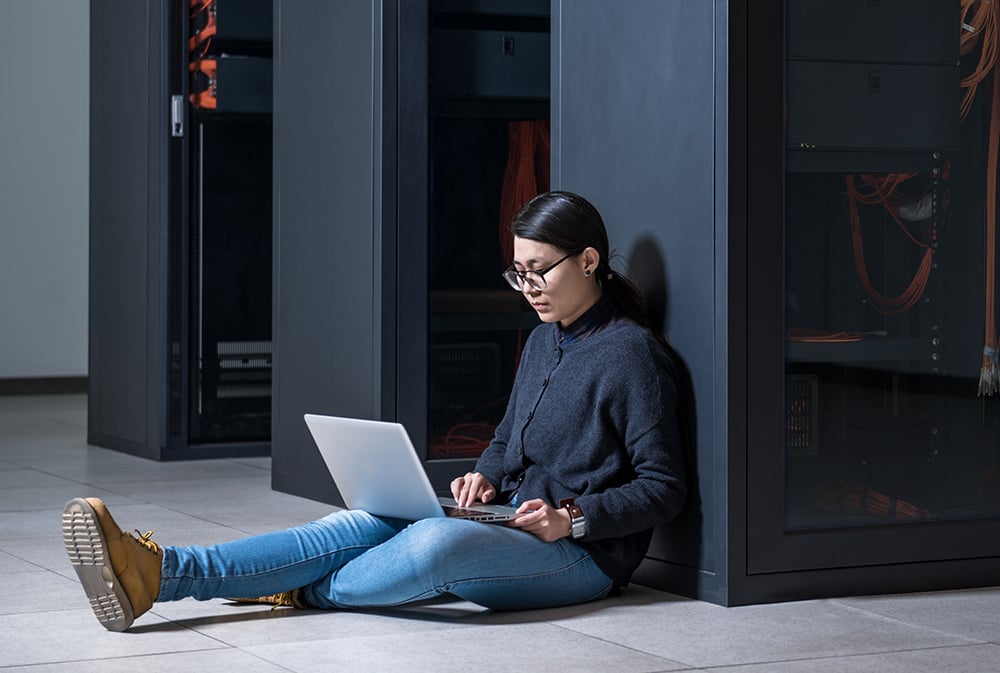 A young woman sitting on the floor against a server cabinet, typing on her laptop.