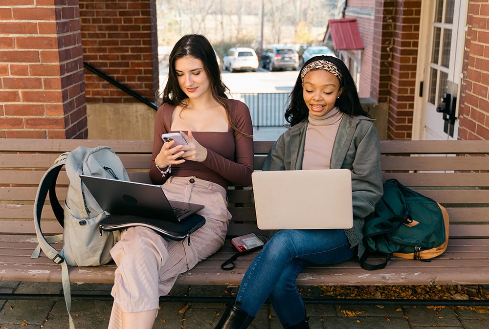 Two colleagues sitting on an outdoor bench, looking at their laptops during conversation.